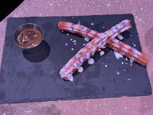 50th Celebration Churro. Churro rolled in cinnamon and strawberry sugar drizzled with marshmallow cream and shimmer sprinkles, and served with chocolate-hazelnut dipping sauce. Available at Frontierland Churro Wagon